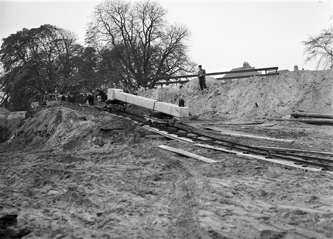 Bouw van het provinciehuis aan de Koningskade. De eerste paal van gewapend beton, een proefpaal, wordt via een werkspoortje getransporteerd naar de heistelling. Tot februari 1959 zullen nog 637 palen volgen.