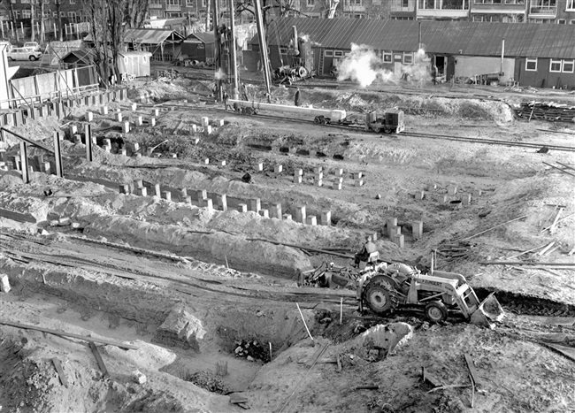 Bouw van het provinciehuis. 508 heipalen worden geslagen voor de fundering van het hoofdgebouw, waarvan de laatste op 27 maart 1964. In dit representatieve gedeelte komen o.a. de Statenzaal en de entreehal. Op de achtergrond is de bouwkeet van de aannemer te zien.