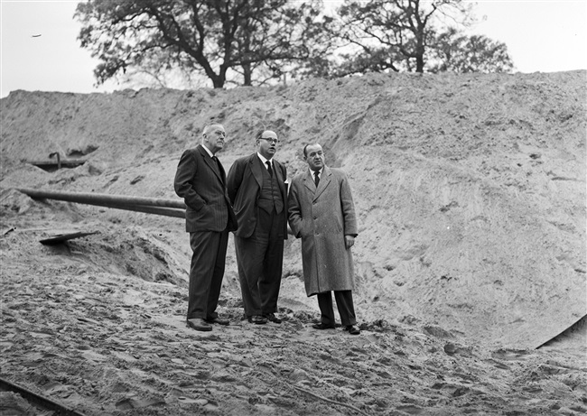 Gedeputeerden kijken naar het heien van de eerste betonpaal van het nieuwe provinciehuis aan de Koningskade. In het midden staat gedeputeerde mr. Harm van Riel.