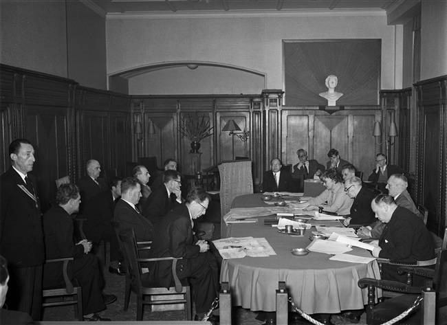 Een vergadering van de Gedeputeerde Staten aan het Korte Voorhout 1. Aan tafel zitten van rechts naar links: gedeputeerde mr. H. van Riel, griffier mr. F.A. Helmstrijd, gedeputeerden dr. J.J.R. Schmal, mevrouw mr. Chr. A. de Ruyter-de Zeeuw en J.P. van Praag.
