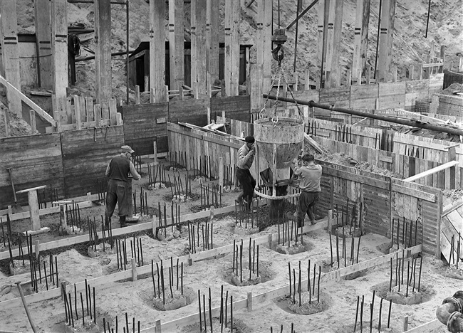 Betonstorters storten met een kubel de vloer van het trappenhuis, van het nieuwe provinciehuis. Op de heipalen staat met streepjes aangegeven waar ze moeten worden weggehakt.