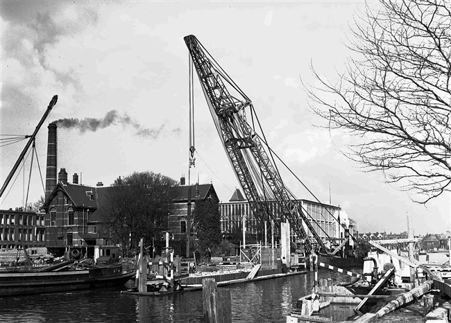 Een stalen damwand wordt geplaatst bij de bouw van de eerste St. Sebastiaansbrug over het Rijn-Schiekanaal. Links het voormalige microbiologielaboratorium van de Polytechnische School.