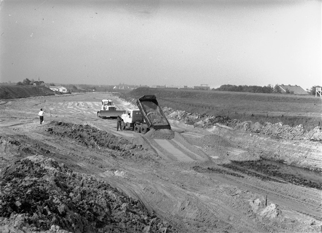 Grondwerken door N.V. B. Smallegange voor de aanleg van de aansluiting van de geplande provinciale weg nr. 4 (tussen Noordwijk en Oegstgeest) in De Klei bij De Krom. Links het dak van café De Viersprong. Rechts het dak van een gebouw van het magazijncomplex van Defensie, De Krom. Op de achtergrond de bebouwing van Katwijk.