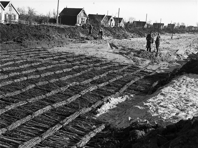 Aanleg van de provinciale weg nr. 29 tussen Gouda en Stolwijk. Om de draagkracht van de bodem te vergroten is een cunet, dat is een brede sleuf, gegraven. Daarin komt een laag houtvezelbalen met daarop een rijzenbed constructie (zie foto), een laag zand en een wegverharding van ipro H-profiel keien.
